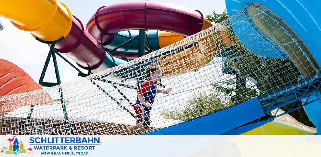 Image of a vibrant waterpark featuring a person climbing through a netted play structure. Multiple colorful water slides spiral in the background against a clear sky, with the logo and location of Schlitterbahn Waterpark in New Braunfels, Texas displayed prominently.