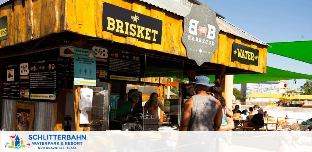 Image shows a bustling BBQ stand at Schlitterbahn Waterpark and Resort in New Braunfels, Texas. Patrons line up at the counter under the shade of an overhang, as staff serve food and drinks. The stand advertises brisket and water, with verdant canopies providing relief from the bright, sunny day. Visitors are casually dressed for a fun day at the park.