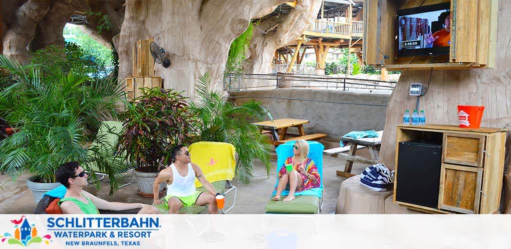 Individuals relax in a shaded cabana area complete with lounge chairs and a TV within the Schlitterbahn Waterpark and Resort, New Braunfels, Texas. Plants add a touch of greenery to the cozy and casual setting, placing emphasis on leisure and comfort.