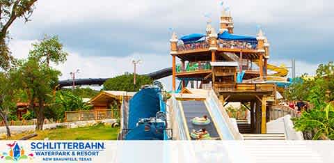 Image shows Schlitterbahn Waterpark and Resort on a cloudy day. A multi-level water slide structure with a blue slide, surrounded by lush greenery, invites visitors. A covered seating area is visible on top, and the enclosed black slide implies thrilling rides await.