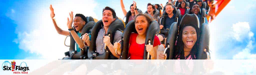 Excited riders with hands in the air on a roller coaster at Six Flags Fiesta Texas, under a clear blue sky. Joy and thrills are evident on their faces. The Six Flags logo is seen on the lower left with 'Fiesta Texas' below it.