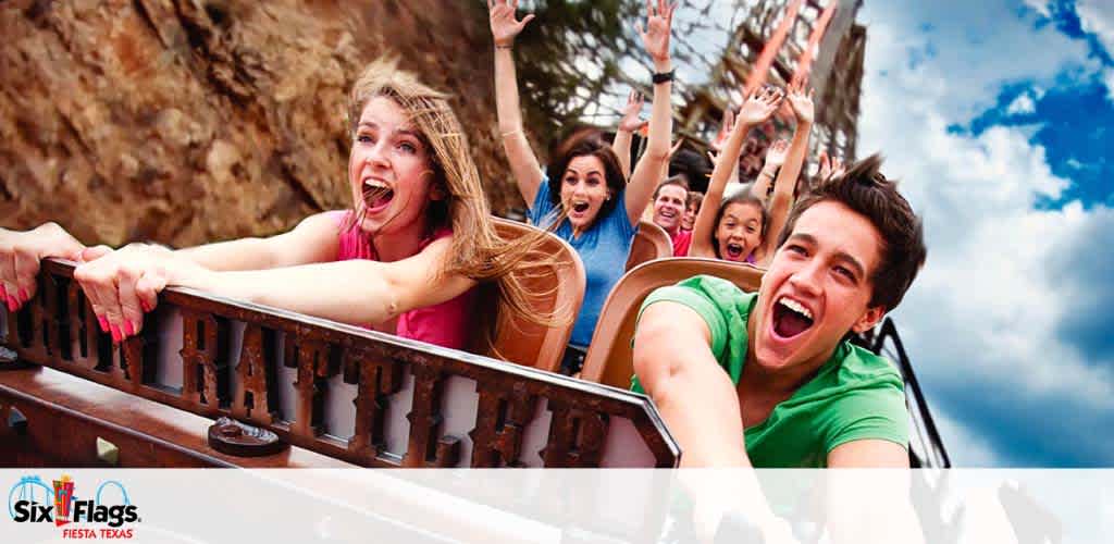 Excited riders on a roller coaster with hands raised in joy, their faces expressing thrill. The backdrop shows a clear sky and rocky landscape, with the Six Flags Fiesta Texas logo at the bottom.