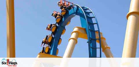 Image shows a blue roller coaster in mid-descent on a track, with its riders raising their hands in excitement. The bright sky serves as the backdrop. Six Flags logo is visible, indicating the coaster is at one of their theme parks.