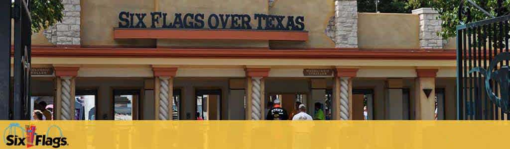 Entrance of Six Flags Over Texas theme park with a sandy beige facade featuring the park's name in capital letters. People are seen walking beneath the brown-trimmed archways of the welcoming structure. A vibrant yellow barrier runs along the bottom of the image.