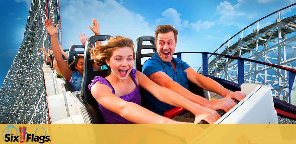 Excited riders on a roller coaster with arms raised. A young woman in purple and a man in blue are in the foreground, smiling broadly against a backdrop of blue sky and coaster tracks. The Six Flags logo is visible in the corner.