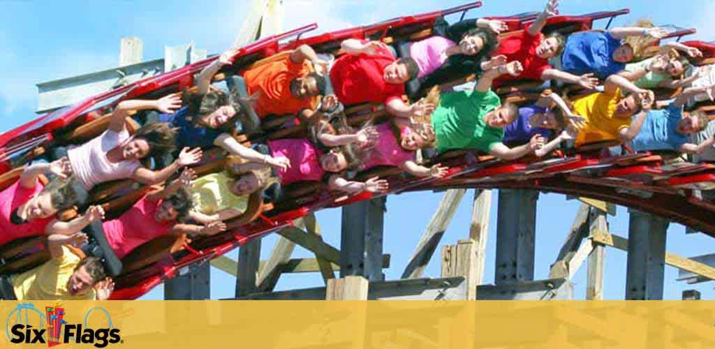 Visitors enjoy a thrilling ride on a red roller coaster at Six Flags, expressing excitement with raised hands. The wooden structure of the coaster track is visible against a clear blue sky. The Six Flags logo adorns the lower left corner.