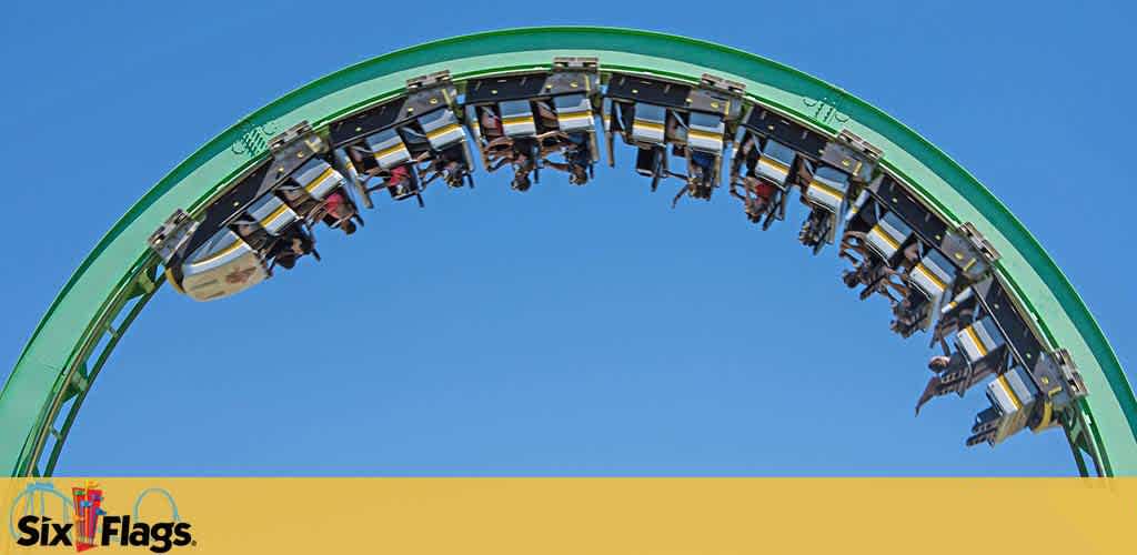 Image features a roller coaster at Six Flags mid-loop with passengers upside down, against a clear blue sky. The Six Flags logo is visible at the bottom left. The coaster is green and yellow, with securely harnessed riders enjoying the thrill.