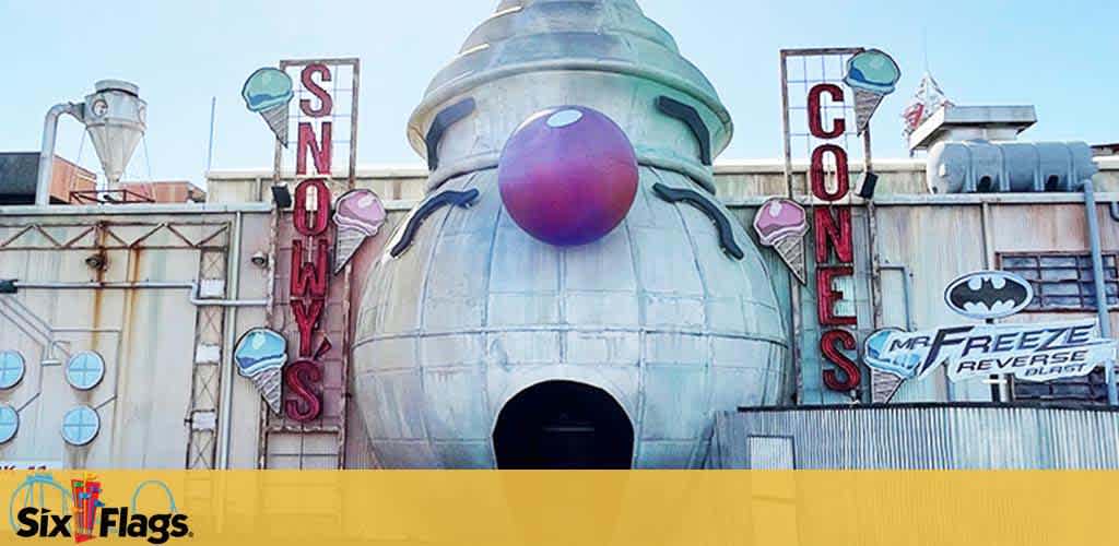 Image of a theme park attraction facade with a large, spherical centerpiece resembling a snowman's head topped with a purple hat. The building has signage reading 'Snowys Cones' in a vertical layout with images of ice cream cones. On the right, a banner for 'Mr. Freeze Reverse Blast' with a Batman symbol is attached to a silvery industrial-looking structure. The Six Flags logo is visible in the lower-left corner, indicating the park's brand.