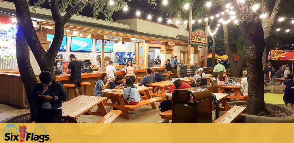 This image shows an outdoor dining area at night with strings of lights above. People are seated at picnic tables, some eating, while others stand by the counter of a food vendor. There are trees interspersed throughout, creating a cozy ambiance. The Six Flags logo is visible, indicating the setting is within an amusement park.