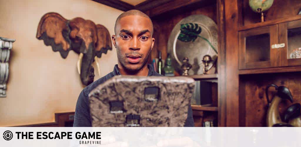 Image shows a person inside a room with a vintage aesthetic, holding a briefcase and looking directly at the camera. Behind them are wooden shelves adorned with various objects, two globes, and a carved elephant head wall mount. The watermark text reads 'THE ESCAPE GAME GRAPEVINE'.