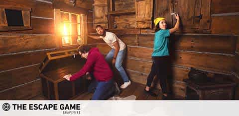 Three people are engaged in an escape room game, actively searching for clues in a wood-paneled room. One individual in a red jacket is examining a piece of paper, another in a white shirt leans forward intently, and the third in a turquoise top reaches upwards. The mood is one of excitement and focused investigation. The Escape Game logo is present, indicating the brand.