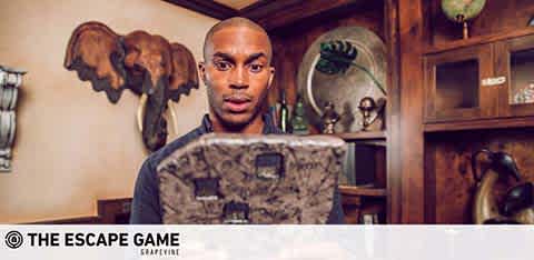 Image depicts a person with a surprised expression holding a large, puzzle-like item. They are in a room with vintage decor, including wooden furniture and wall-mounted animal sculptures. The logo 'THE ESCAPE GAME GRAPEVINE' is visible.