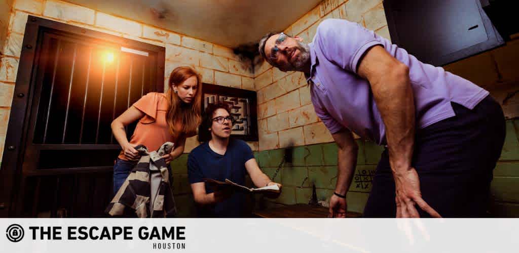 Three participants are engrossed in an escape room challenge at The Escape Game Houston. They're attentively searching for clues in a dimly-lit, prison-themed room, with one of them bending over to inspect something closely, another looking on with a clipboard in hand, and the third kneeling and examining a wall. The excitement of the game is evident on their focused faces.