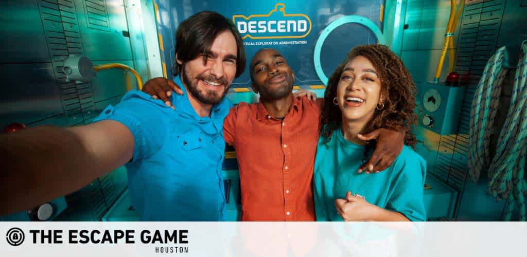 Three joyful friends are taking a selfie together inside 'The Escape Game' room in Houston, with a theme of 'DESCEND' indicated in the background. The two men and one woman display wide smiles, reflecting a fun moment shared. Branding for 'The Escape Game' is visible.