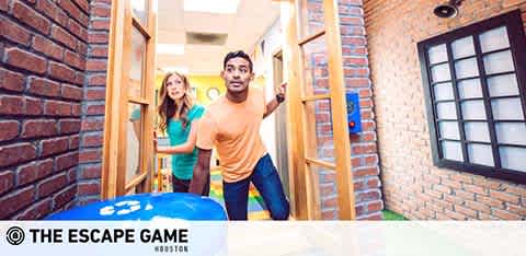 Image shows a man and woman emerging enthusiastically from a doorway in a brick-walled corridor with escape room branding. The man in an orange shirt points forward, while the woman in the background expresses surprise. A blue object with symbols is in the foreground. The Escape Game logo is visible.