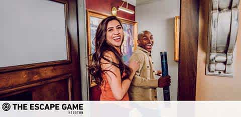 Two people are laughing joyfully as they exit an escape room game. The person in the front, with long hair, is looking back, laughing, while the person behind is holding a blue clue file, smiling widely. Both seem ecstatic, likely having solved the game. A logo with 'THE ESCAPE GAME' is visible on the bottom right.