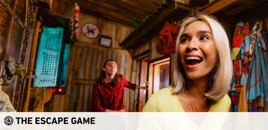 Photo taken inside The Escape Game venue, showing a wood-paneled room with vintage decor. A woman in the foreground expresses excitement with eyes wide and a joyful smile. A man in a red flannel shirt stands in the background, focusing on a part of the room with an observant gaze. The atmosphere is lively, suggesting fun and adventure.