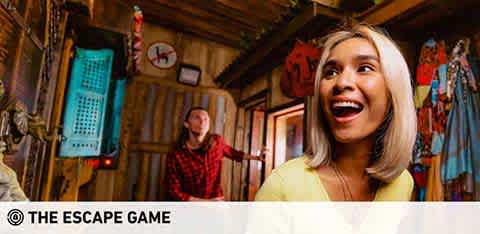 Excited woman in the foreground at an escape room, with a joyful expression, looking upwards. A man in the background actively engages with the room's puzzle elements. The room is adorned with colorful, eclectic decor and various objects, creating an immersive setting for the game. Clock on the wall indicates game time. Logo: The Escape Game.