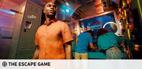 Image of a man standing confidently in a vibrant escape room setting, looking to the side with anticipation. In the background, a group of people is actively engaging with the room's puzzle elements by a control station. The environment hints at an immersive and challenging adventure experience, with © THE ESCAPE GAME visibly branded.