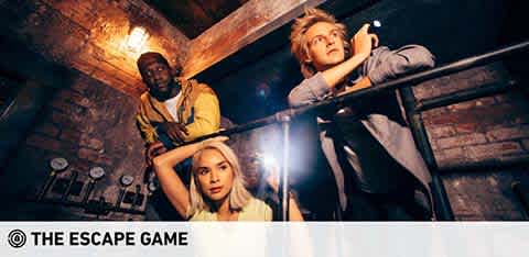 Image shows three people engaged in an escape room activity, looking around with flashlights for clues. The setting has a rustic aesthetic with brick walls, indicative of a mystery or adventure-themed game. Text at the bottom of the image reads 'THE ESCAPE GAME'.