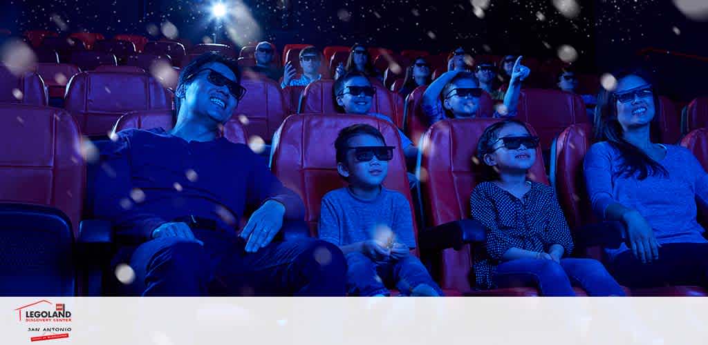 Audience enjoying a 4D cinema experience at LEGOLAND in San Antonio. They are wearing 3D glasses and smiling in a dimly lit theater with blue lighting and onscreen effects that appear to float around them.