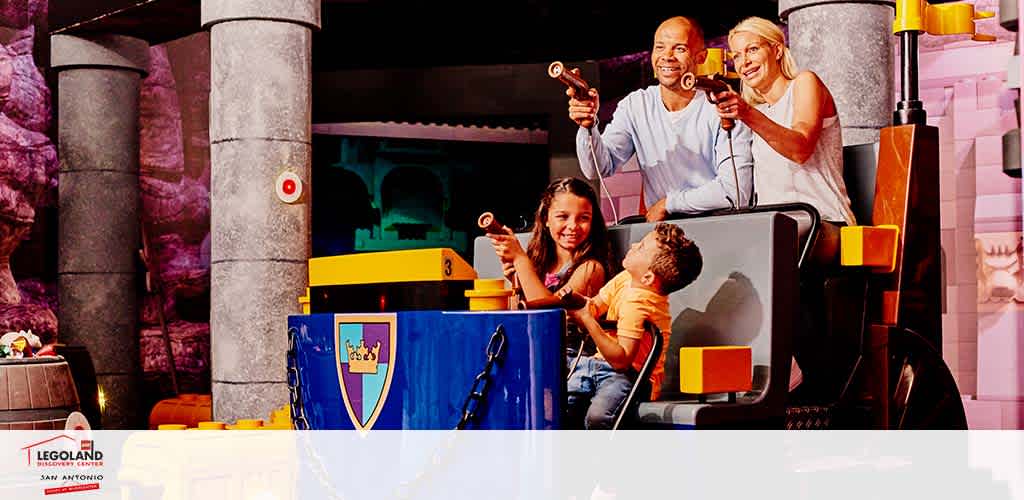 A joyful family of four is riding an interactive ride at LEGOLAND San Antonio. They are seated in a blue cart adorned with a crest, embarking on an adventure. The backdrop features whimsical, LEGO-inspired stone columns and vibrant scenery. The parents are holding up wands, engaging with the ride, as their daughter smiles widely and their young son looks ahead with excitement.