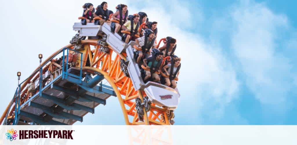 Visitors enjoy a thrilling ride on a roller coaster at Hersheypark. The coaster, with an orange track, is captured mid-descent as excitement is visible on the riders' faces. Clear skies backdrop the scene with the park's logo on the bottom left.