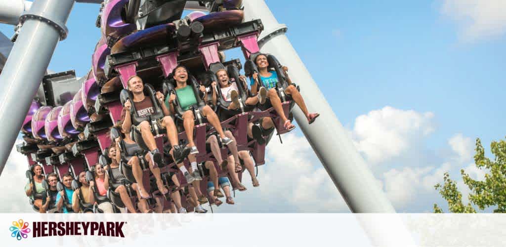 Visitors enjoy a roller coaster ride at Hersheypark on a sunny day. They are strapped into a purple-trimmed coaster with legs dangling freely, showing expressions of thrill and joy. Trees and a clear blue sky with some clouds form the background. The park's logo is visible in the corner.