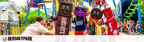 Visitors pose with life-sized, mascot versions of Hershey's chocolate bars at Hersheypark. The characters, in various colors, greet guests near a vibrant roller coaster under a clear sky. The scene is full of energy and excitement, welcoming all to a day of amusement.