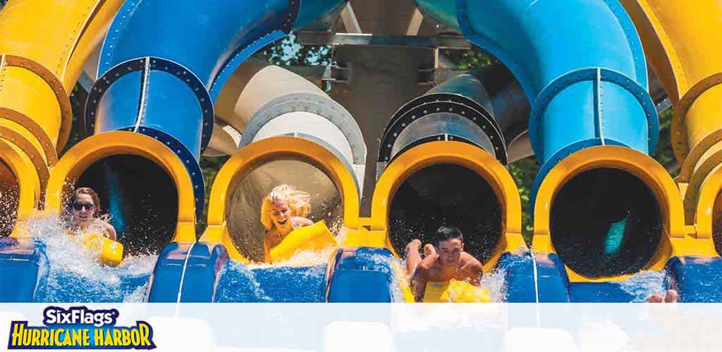 Excitement unfolds at a water park where visitors splash down vibrant blue and yellow water slides. Water sprays as they emerge, smiling and enjoying the thrill. In the background, more slides promise continued fun under a sunny sky. The logo of Six Flags Hurricane Harbor adorns the image, suggesting a lively aquatic adventure.