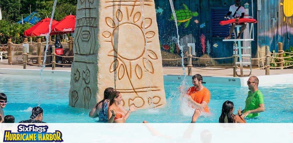 Guests enjoy a sunny day at Six Flags Hurricane Harbor. People are seen splashing in a shallow pool near a large decorative pillar with a sun motif. A lifeguard watches over from a nearby stand as colorful slides await in the background underneath blue skies and umbrellas dot the deck area.