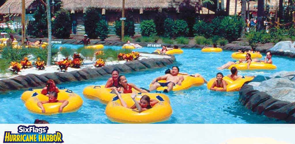 Visitors are enjoying a leisurely float on a lazy river ride at Six Flags Hurricane Harbor. They're in bright yellow inner tubes, drifting along a blue water channel surrounded by artificial rock formations and lush greenery. The atmosphere is tropical, with straw-covered pavilions in the background.
