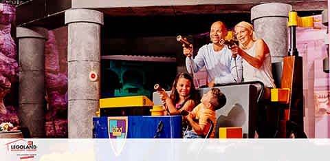 Image shows a family enjoying a ride at LEGOLAND. Two children and two adults are seen waving and smiling on a colorful ride vehicle with LEGO-inspired designs, passing under a stone-like archway, adding fun to the vibrant amusement park atmosphere.