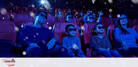 Image of a family sitting in a movie theater at LEGOLAND, wearing 3D glasses. They are smiling and looking forward at the screen, surrounded by a dim blue light and floating white particles, suggesting a 3D movie experience.