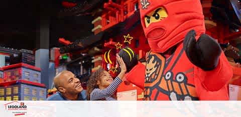 Image of a joyful child raising her hand towards a larger-than-life LEGO Ninjago character mascot, with an adult smiling beside her. The scene is set against a vibrant LEGO themed backdrop at LEGOLAND, invoking a sense of excitement and fun.