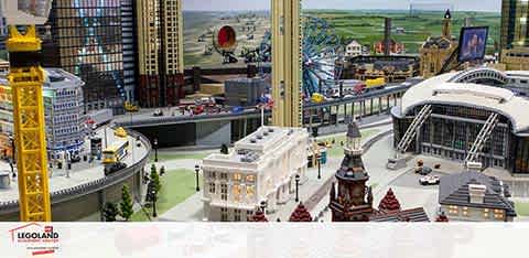 This image shows a vibrant Lego cityscape at LEGOLAND. It features detailed Lego constructions of skyscrapers, various buildings, and a large stadium. The scene includes city streets bustling with miniature Lego cars and includes lush green spaces. The Legoland watermark is visible at the bottom.