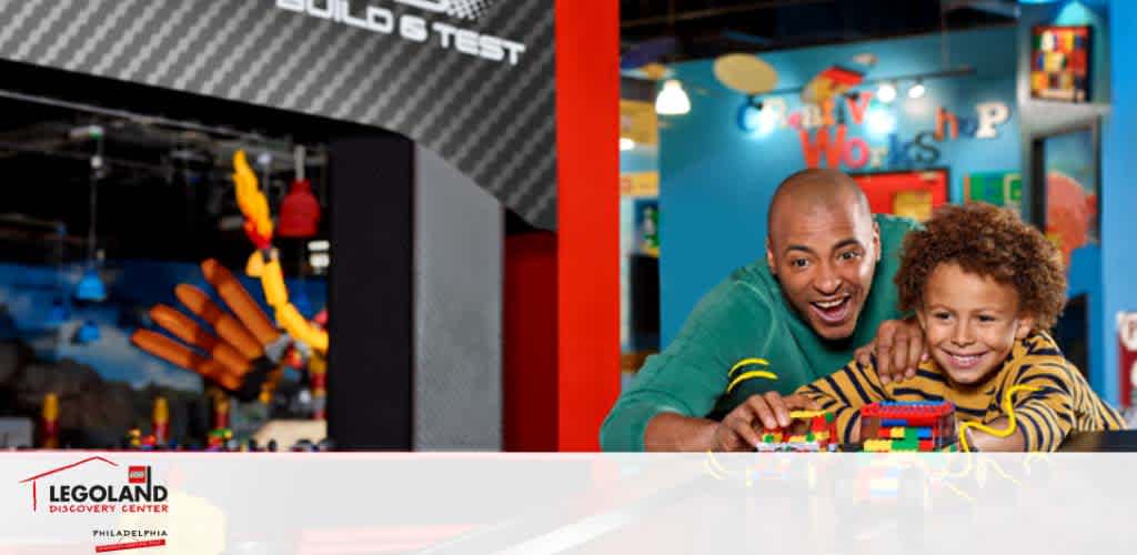 Image of a joyful bald man and a child with curly hair at LEGOLAND Discovery Center. They are smiling and posing with a colorful Lego creation in a vibrant room filled with Lego sets.  Build & Test  sign and  Creative Workshop  in view.