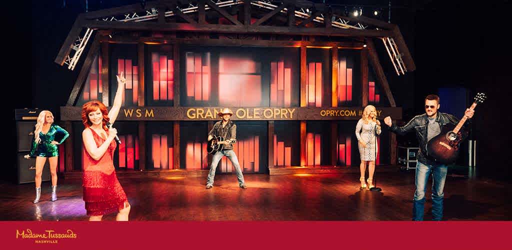 Image of a stage featuring four lifelike wax figures resembling country music performers, with the Grand Ole Opry backdrop. Each figure is styled distinctly, as if captured mid-performance with costumes and musical instruments. The setting conveys a celebratory showcase of country icons.