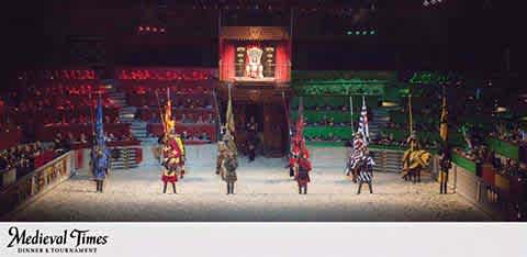 Image shows an indoor Medieval Times jousting arena. Performers in colorful medieval costumes stand on the sandy floor. Spectators are seated in color-coded sections around the arena. A red and yellow banner with the Medieval Times logo is in view.