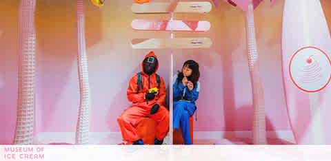 Two individuals are seated on a pink bench in an ice cream-themed setting, marked by playful oversized decorations. The person on the left is wearing an orange jumpsuit and sunglasses, holding a camera, while the one on the right dons a blue jumpsuit, enjoying an ice cream cone. A series of large faux ice cream scoops hang above them, with soft pink walls encasing the whimsical scene.