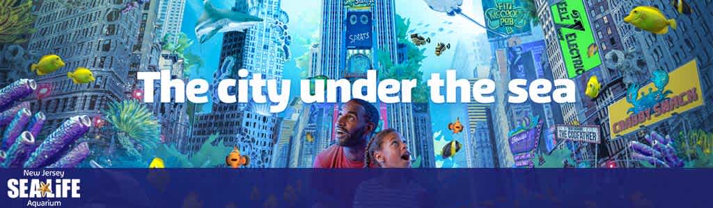 Promotional banner featuring the slogan 'The city under the sea' for New Jersey SEA LIFE Aquarium. A man and a child look up in awe at a vibrant underwater scene blending city architecture with marine life. Brightly colored fish swim among coral and skyscrapers.