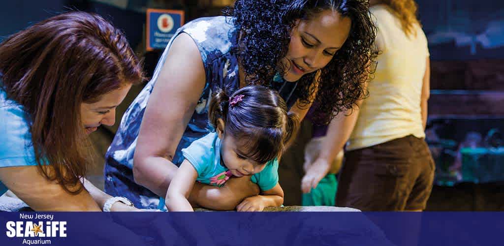 Image displays three individuals, two adults and a child, engaged in an activity at the New Jersey SEA LIFE Aquarium. The woman on the left leans over with a smiling toddler, while the woman on the right assists, both showing the child something of interest. The background suggests an underwater theme. The aquarium logo is visible in the corner.