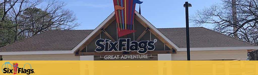 Image depicts the entrance of 'Six Flags Great Adventure' theme park. The park's name is prominently featured on the facade in bold letters, with a decorative logo of a fluttering flag above. The background shows a clear sky and trees, with a glimpse of a roller coaster structure seen on the right.