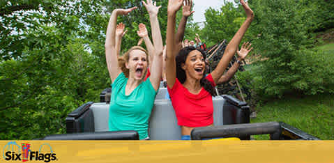 Image shows two people on a roller coaster, their arms raised in excitement. One person wears a green shirt, the other red. They appear to be shouting joyfully. In the foreground is a bright yellow safety bar. Lush greenery is in the background. The Six Flags logo is visible.