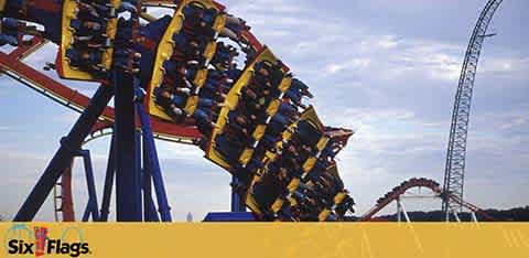 Image of a roller coaster from Six Flags with a clear sky in the background. The coaster has yellow tracks and blue supports. Thrill-seekers in secure seats with over-the-shoulder restraints are mid-ride, appearing to be experiencing a gravity-defying moment. The Six Flags logo is visible in the bottom left corner.