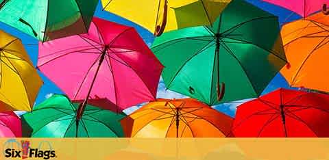 This image presents a vibrant array of open umbrellas in assorted colors including pink, green, blue, yellow, and red. They appear to be suspended against a clear blue sky. In the lower left corner, the logo for 'Six Flags' theme park is visible.