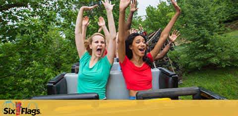 Image shows two people expressing excitement with their arms raised while riding a roller coaster. One is wearing a blue shirt and the other red. Trees in the background indicate an outdoor setting. The Six Flags logo is visible, suggesting the location is a theme park.