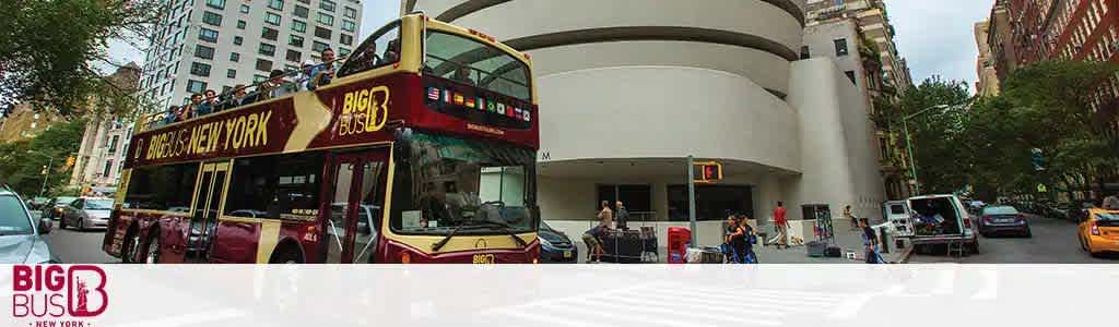 Image displays a red double-decker tour bus labeled Big Bus New York in front of a modern curved white building. Several vehicles and pedestrians are visible on the street, along with some green trees lining the sidewalk. A clear sky encompasses the background.