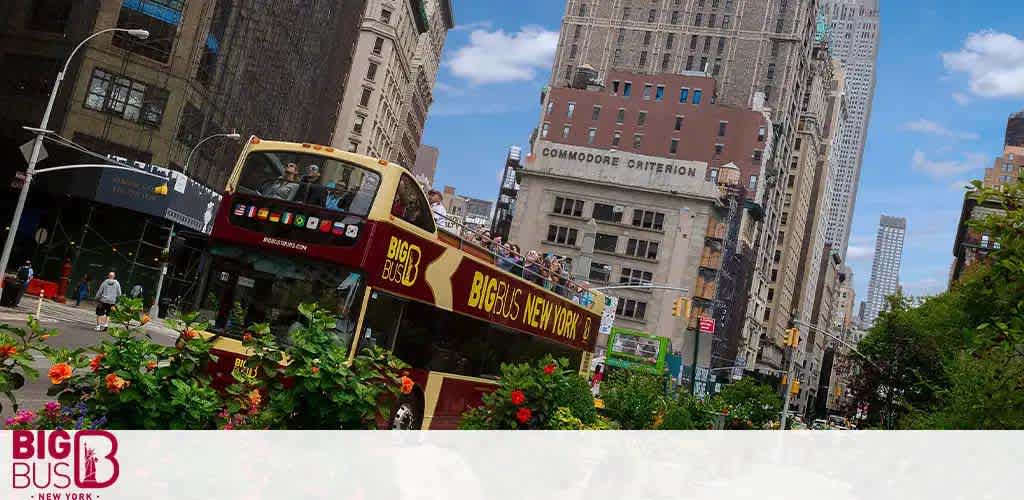 Image of a red, open-top sightseeing bus labeled 'Big Bus New York' with passengers on the upper deck, cruising through a sunny, bustling city street with buildings and a clear blue sky above. Foreground shows vibrant flowers, hinting at a city garden or park.