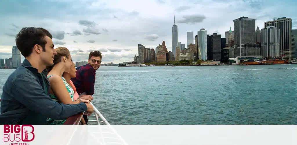 Three people are seen enjoying a boat ride with a view of New York City's skyline. The nearest person smiles at the camera, while the pair gazes out at the urban backdrop, featuring the iconic One World Trade Center. The water is calm, with scattered clouds overhead. The image includes the Big Bus New York logo.